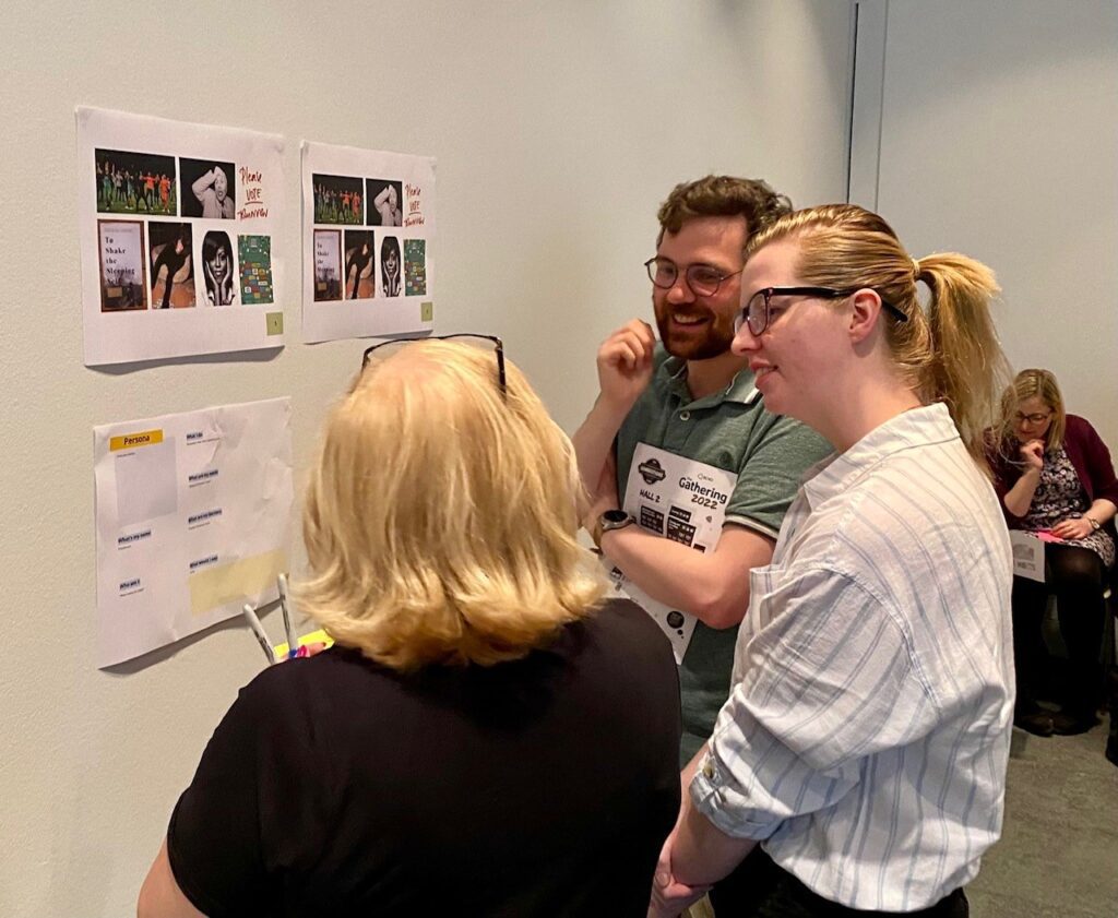 A group of 3 people standing in a room looking at two sets of 6 photographs on a wall. The photos are taken from social media accounts and the people are creating a persona about that person based on the photos they post